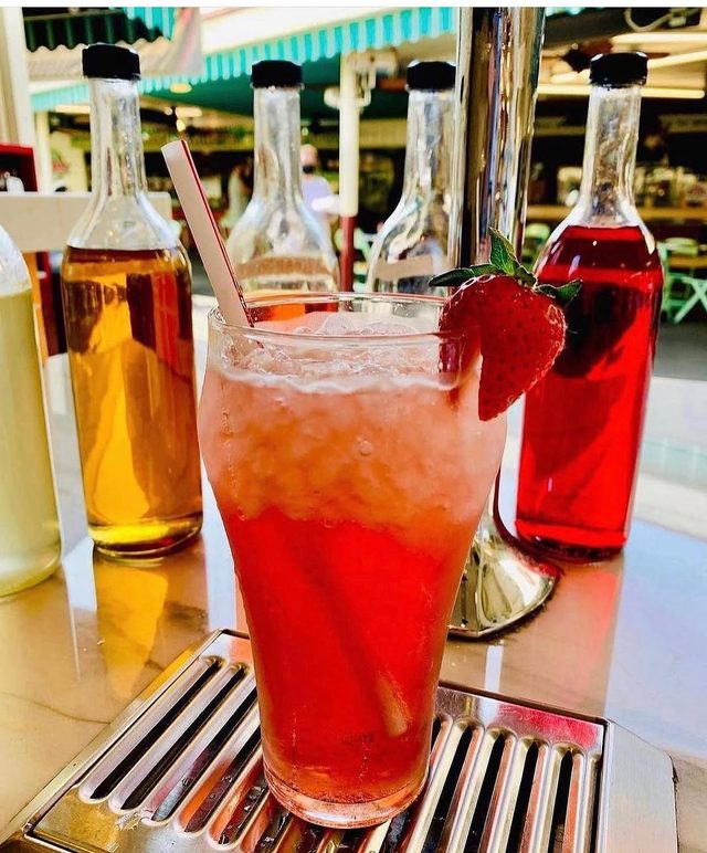 A tall glass of red Italian soda, the coloured syrup bottles lined up behind.