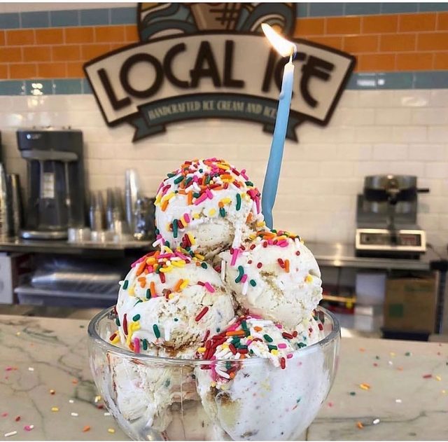 A coupe of scoops covered in rainbow sprinkles, under the Local Ice sign.