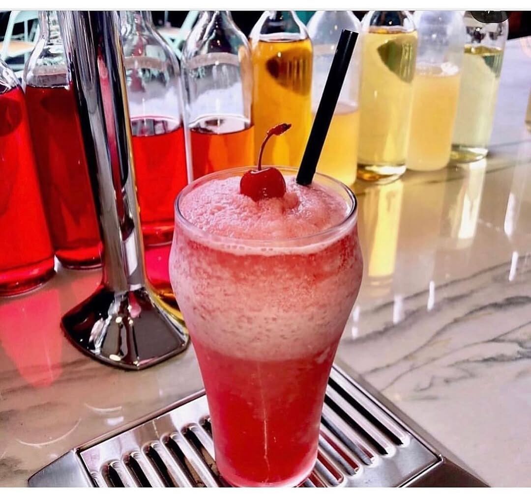 A tall glass of raspberry-red soda topped with foam and a cherry, on the marble fountain, with the coloured syrup bottles lined up behind.