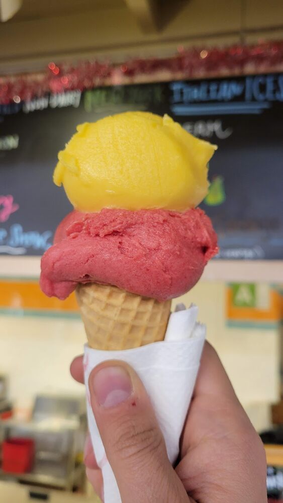 A waffle cone with a yellow scoop and a raspberry scoop, in front of the Italian ice sign.
