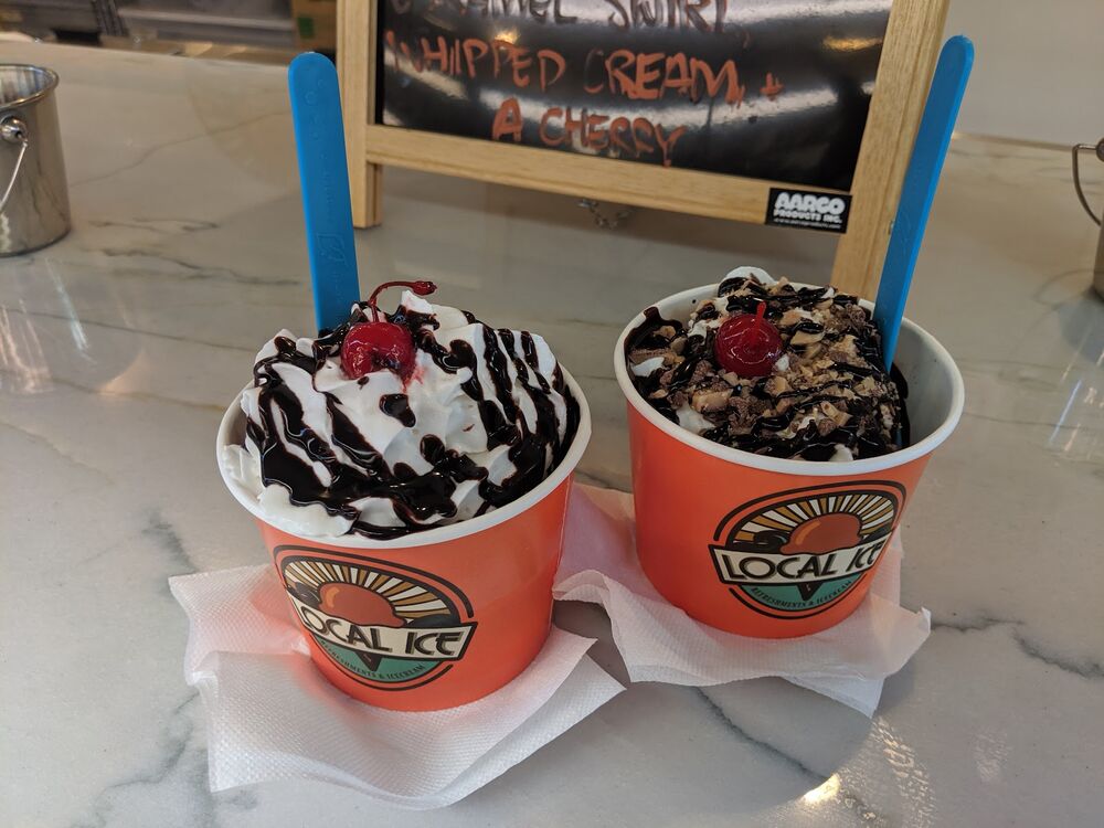 Two Local Ice cups on the marble counter, whipped cream, chocolate sauce and a cherry on each.